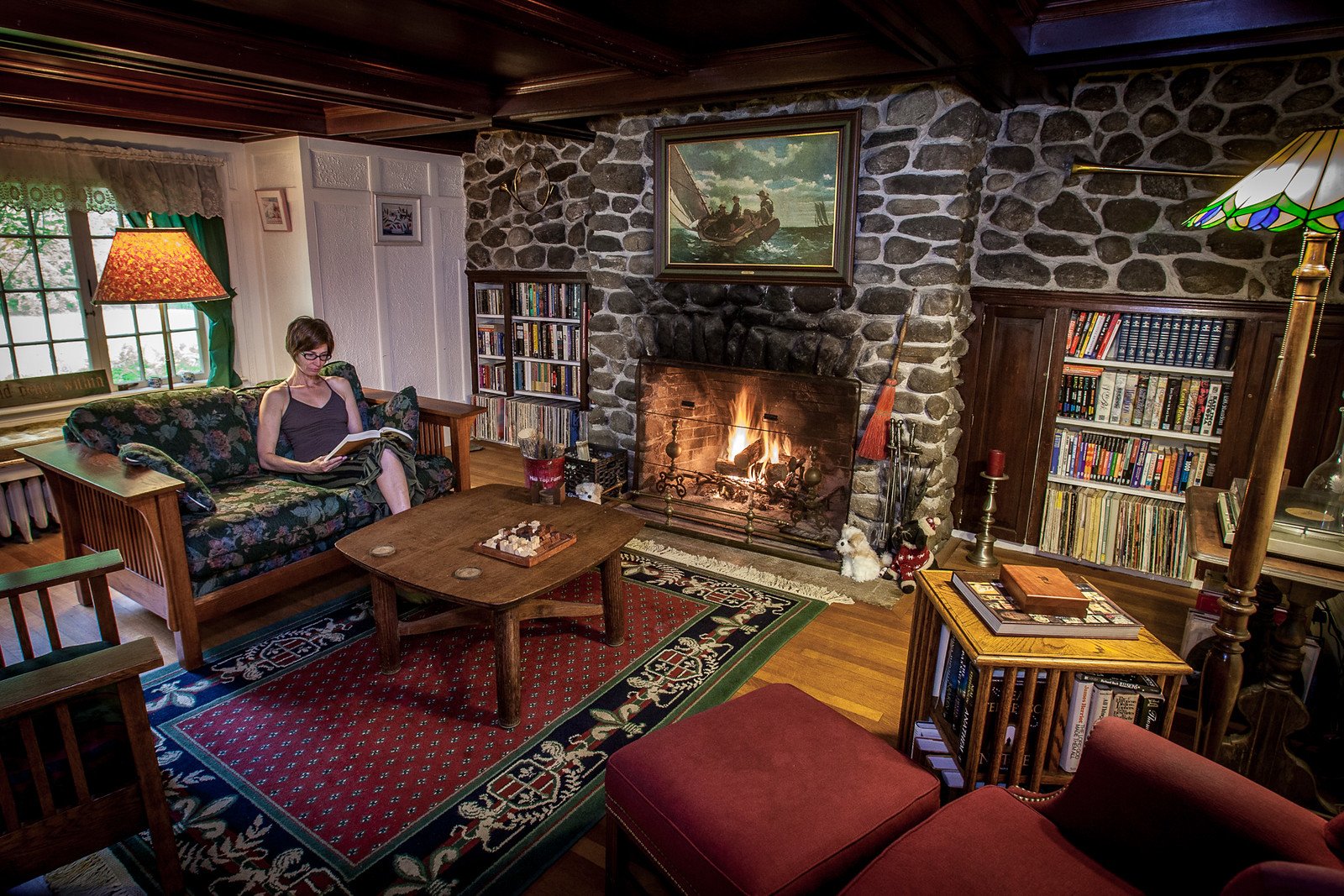 A guest reads on a floral sofa beside a lit stone fireplace in the inn's wood-paneled common room, with built-in bookshelves and a sailboat painting above the mantel.