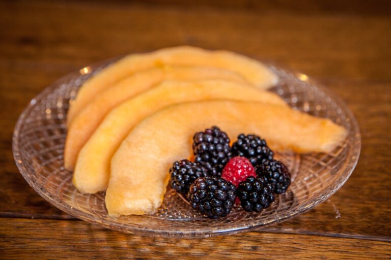 Sliced cantaloupe fanned on a glass plate with fresh blackberries and a raspberry.