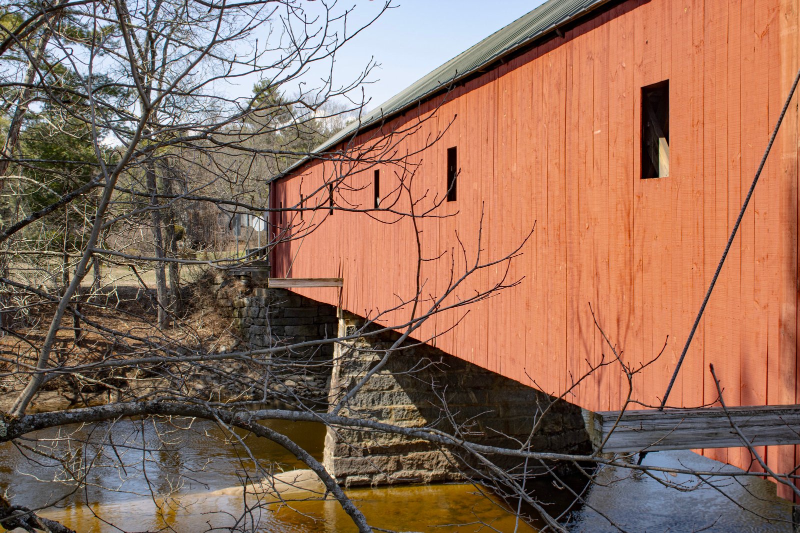 A red New England covered bridge crossing a stream, with bare winter trees and stone footings, in Swanzey, New Hampshire.