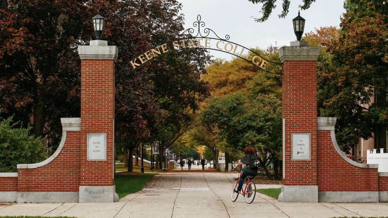 The brick entrance gate to Keene State College in autumn, with a cyclist riding beneath the archway and trees in fall color.
