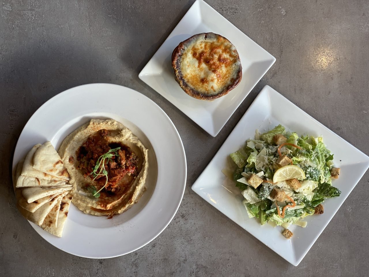 A shared appetizer spread of hummus with pita, French onion soup, and a Caesar salad on a restaurant table.