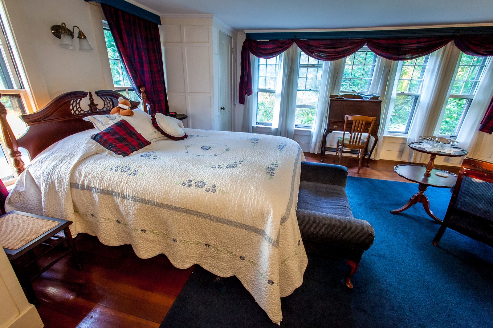 A four-poster bed with a white quilted spread and a red-and-navy tartan pillow in the Manor Room, under matching plaid drapery.