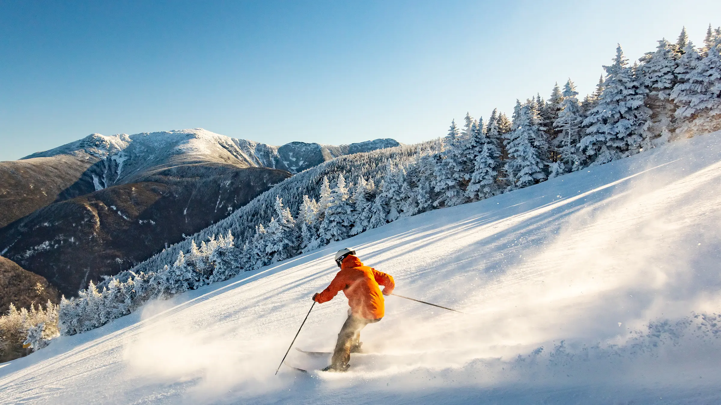 A skier in an orange jacket carves through fresh powder on a sunny New Hampshire mountain trail, framed by snow-covered evergreens.
