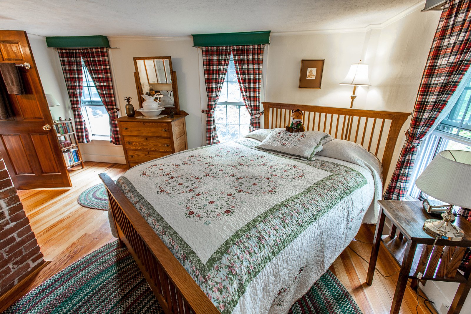 The Stewart Room's Queen bed with a white, green, and pink floral quilt, beside an exposed brick chimney and plaid curtains.